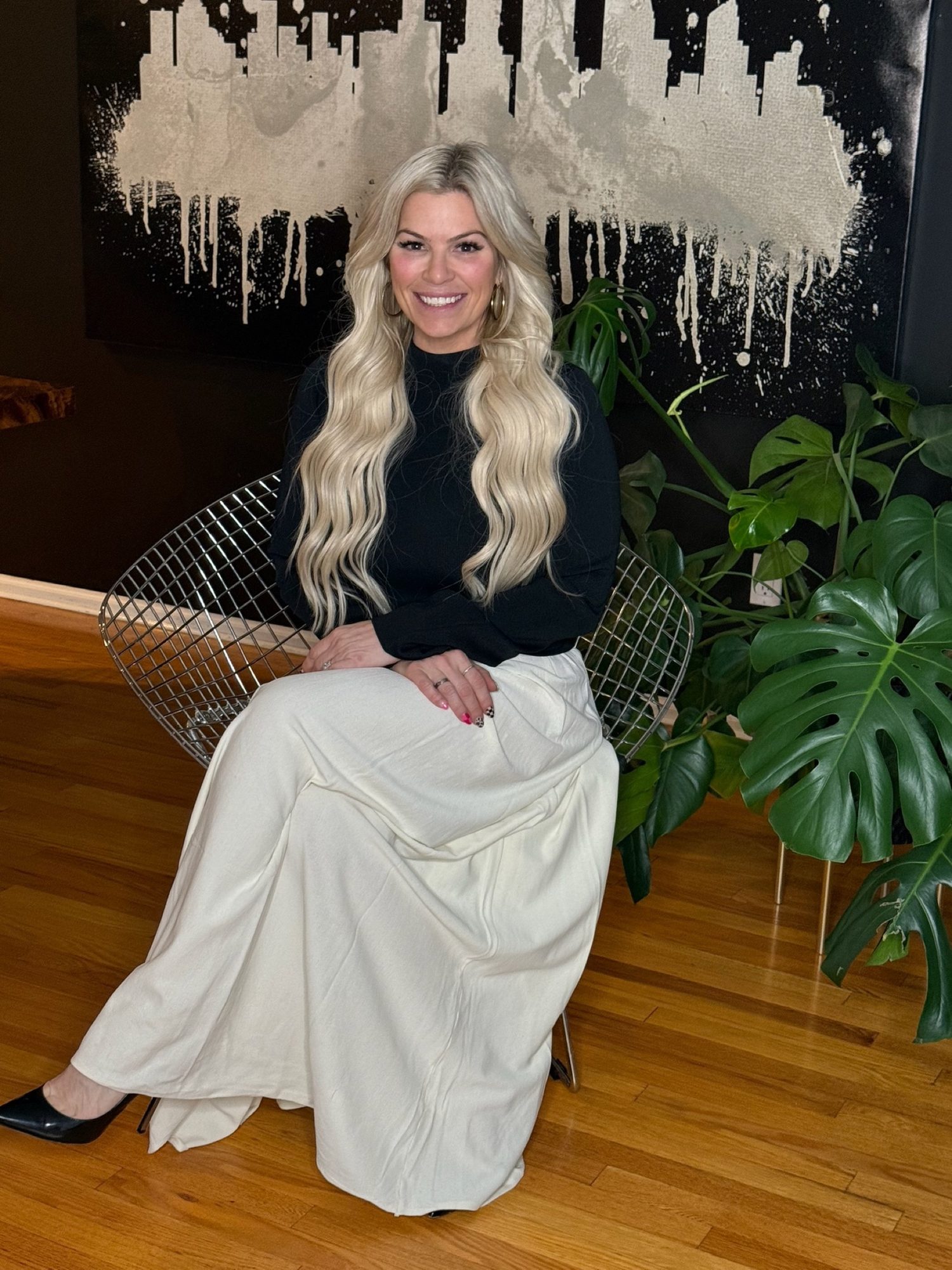 Dr. Christine Hanczaryk seated in a white skirt beneath the drip painting, a monstera beside her