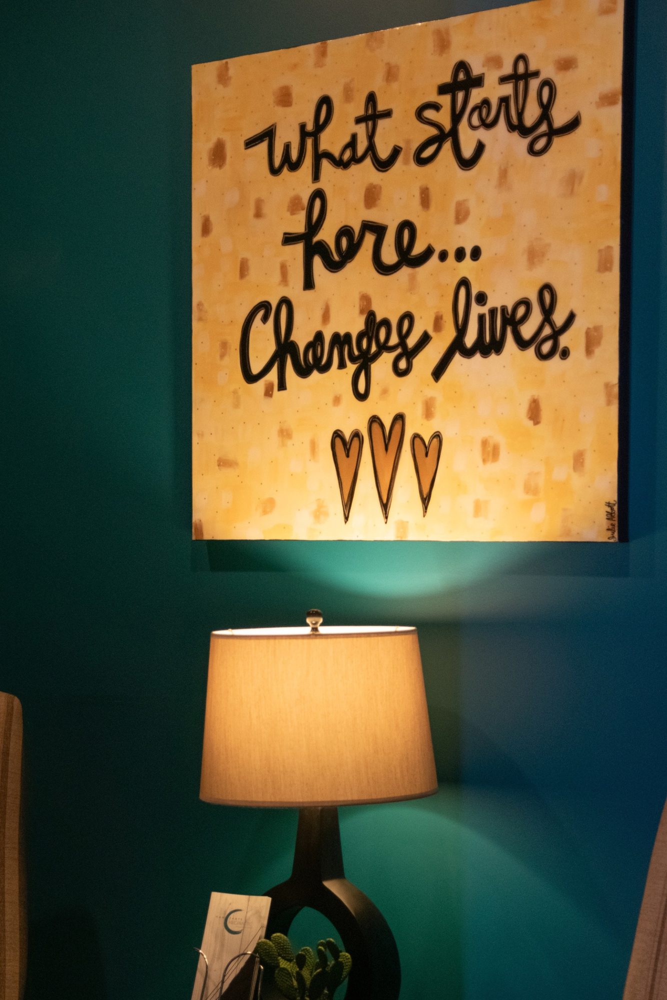 A hand-lettered canvas that reads What starts here changes lives, above a table lamp